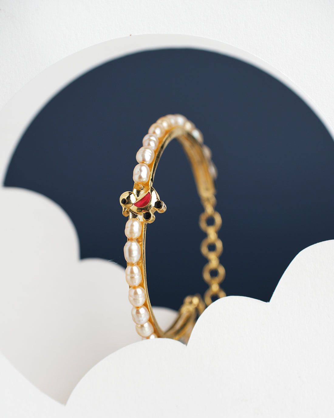 The Little Duck Pearl Bangle for kids by Chandrani Pearls India features a gold design with white pearls and a small red and gold accent, set against a dark background with white curved shapes in the foreground.