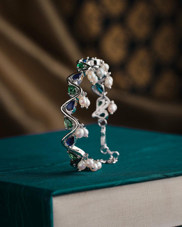 The Zigzag Cluster Pearl Bangle from Chandrani Pearls, featuring a design with pearls and gemstones in green and blue hues, rests on a teal fabric-covered book.