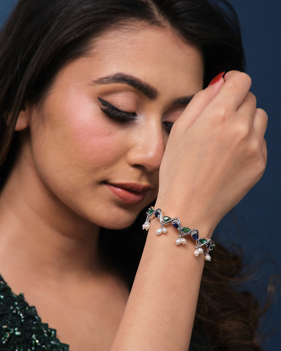 The Zigzag Cluster Pearl Bangle from Chandrani Pearls, featuring a design with pearls and gemstones in green and blue hues, rests on a teal fabric-covered book.