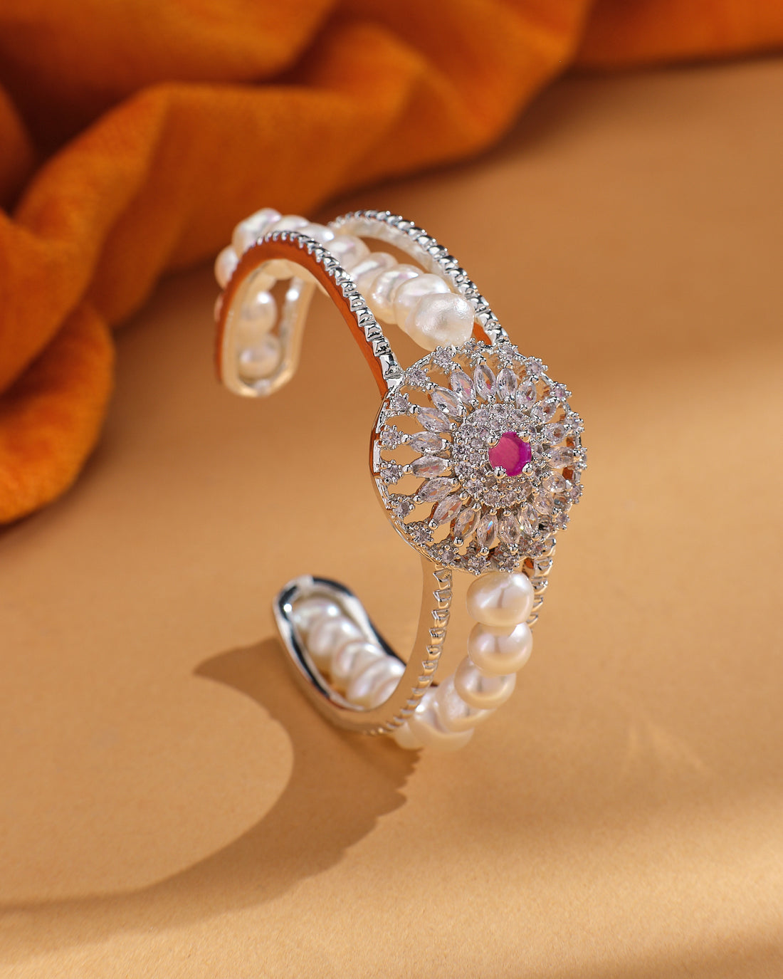 The Gorgeous & Beautiful Shine with Red CZ Bangle by Chandrani Pearls features a floral design with CZ stones, pearls, and a striking red stone, elegantly displayed on beige with orange fabric in the background.