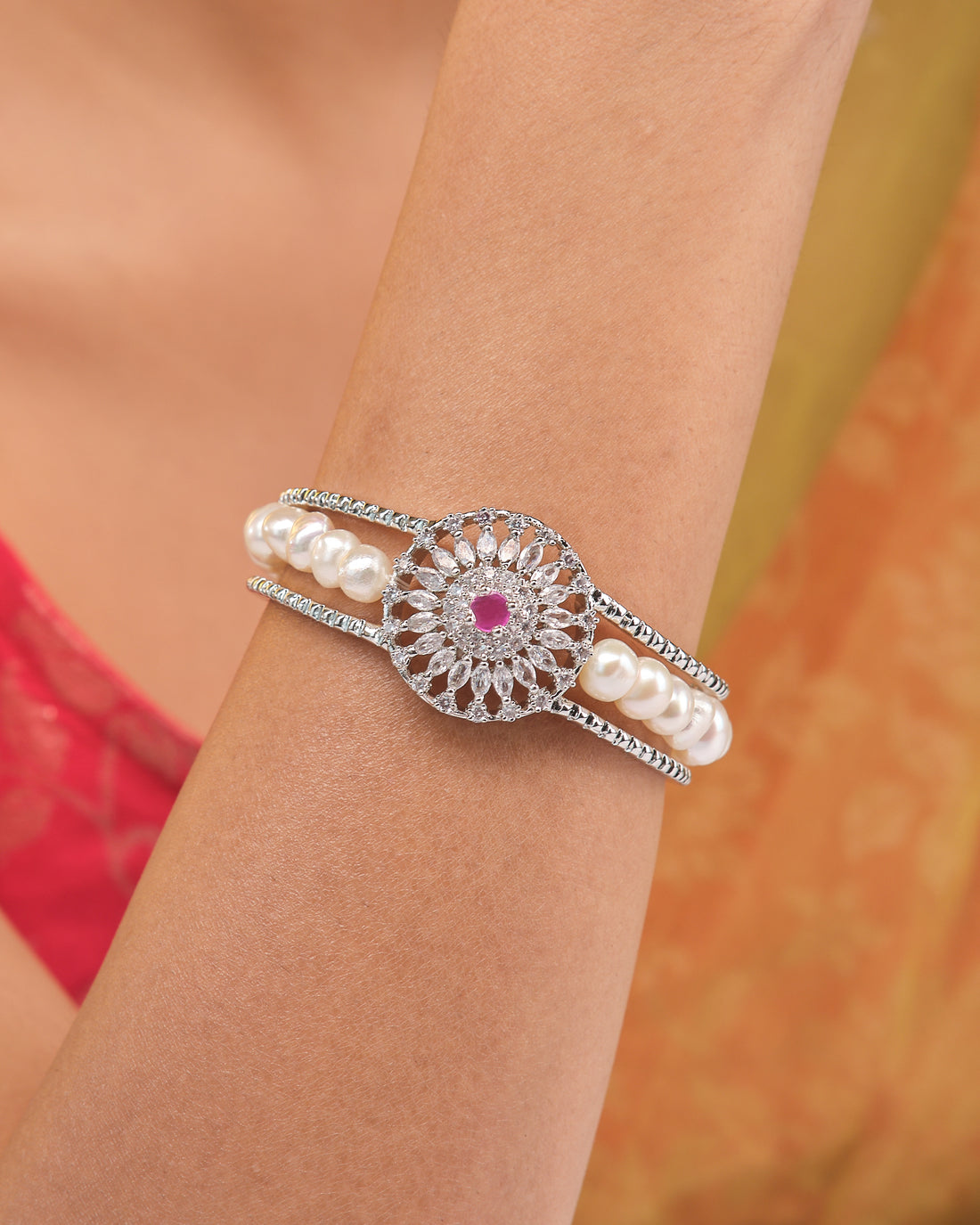 The Gorgeous & Beautiful Shine with Red CZ Bangle by Chandrani Pearls features a floral design with CZ stones, pearls, and a striking red stone, elegantly displayed on beige with orange fabric in the background.