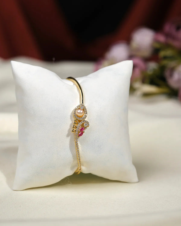 The Joella CZ Stone Bangle by Chandrani Pearls India features an elegant curved design with sparkling CZ stones, pearls, and a pink accent, presented on a white cushion for a glamorous display.