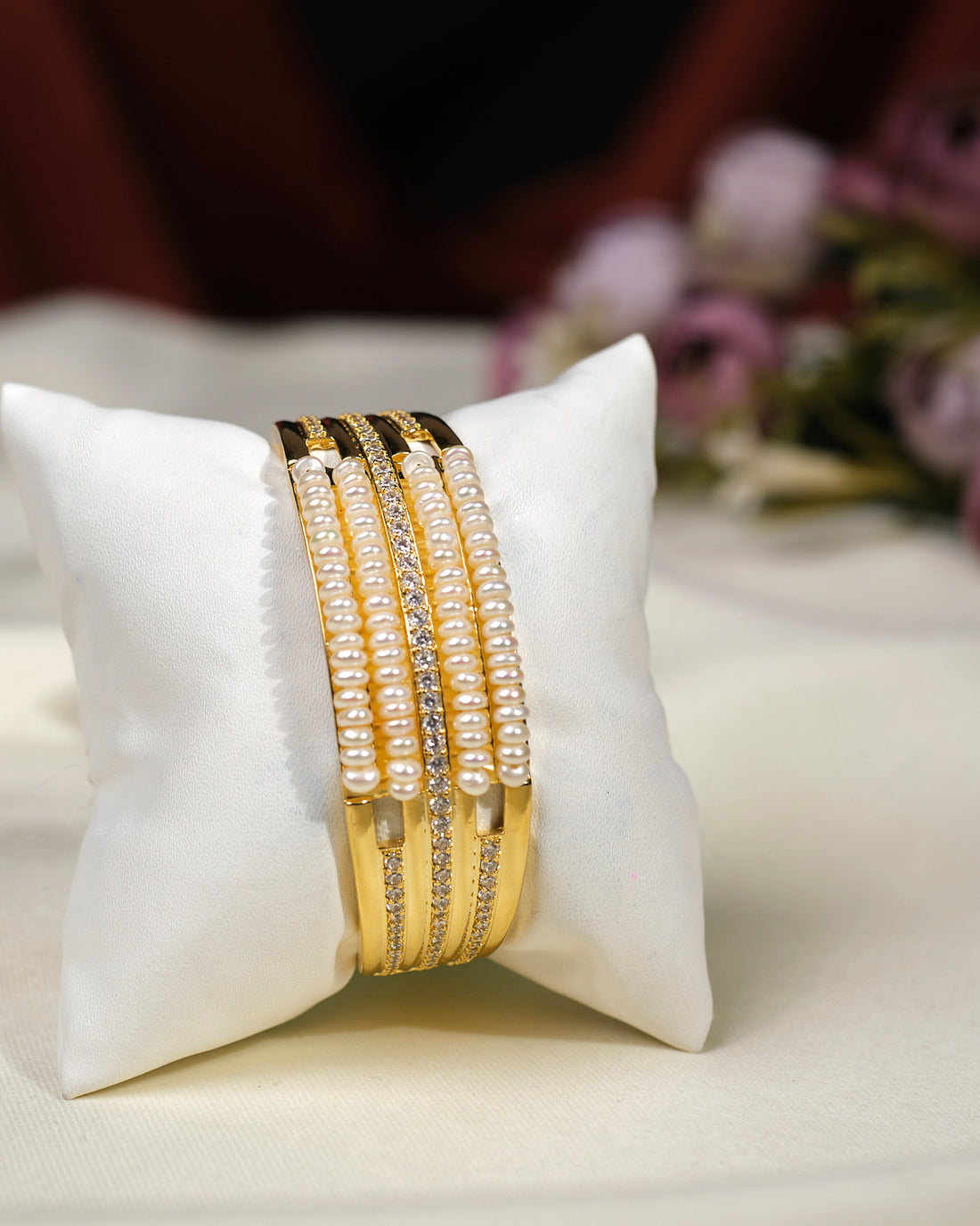 The Natalie CZ Stone Bangle by Chandrani Pearls India features an elegant gold design with pearls and crystals, displayed on a white cushion with blurred purple flowers in the background.