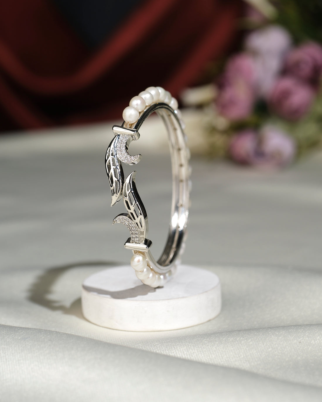 The Leafy Pearl Bangle by Chandrani Pearls India, featuring pearl accents and detailed metalwork, is displayed upright on a round white stand with blurred flowers in the background.