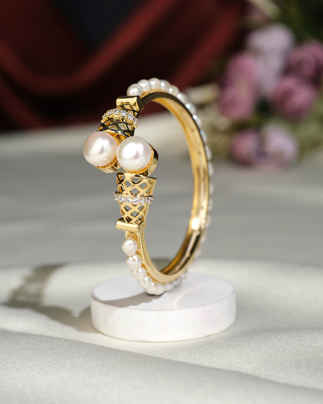 The Trendy White Bangle by Chandrani Pearls India is displayed upright on a white circular stand, set against a blurred floral background.