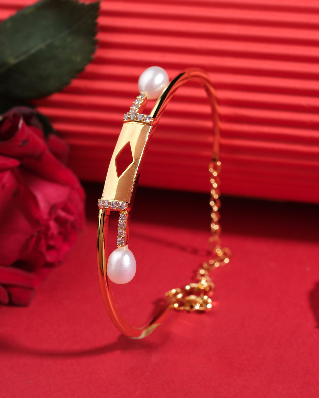 The Quad and Pearl 925 Silver Bangles by Chandrani Pearls India feature a geometric centerpiece, pearls, and a chain clasp, elegantly displayed on a red surface beside a red rose.
