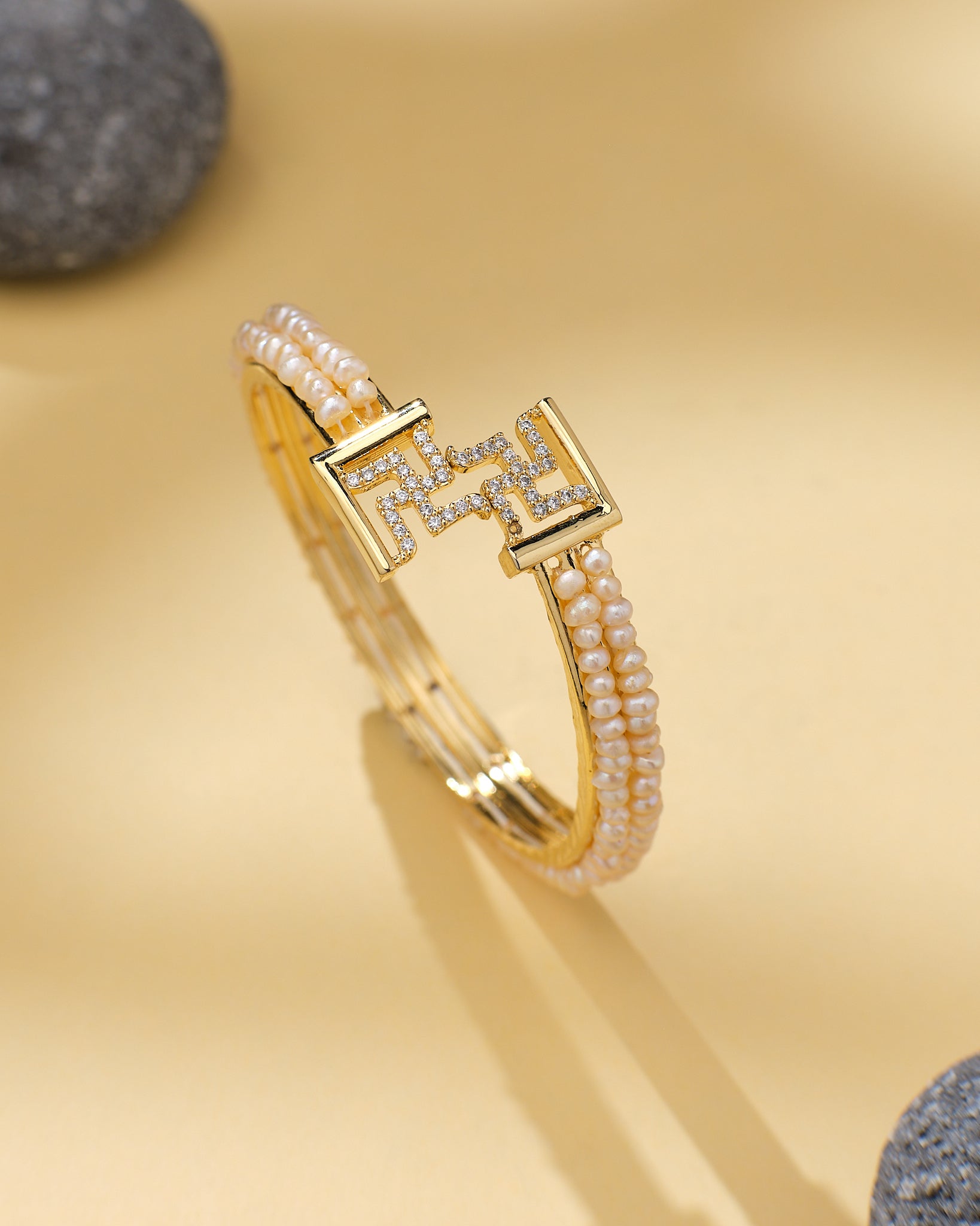 Swasthik White Pearl & CZ Cuff Bangle by Chandrani Pearls—a chic statement piece with two rows of small pearls, CZ stones, and a geometric clasp, styled on a light beige surface with smooth gray stones in the background.
