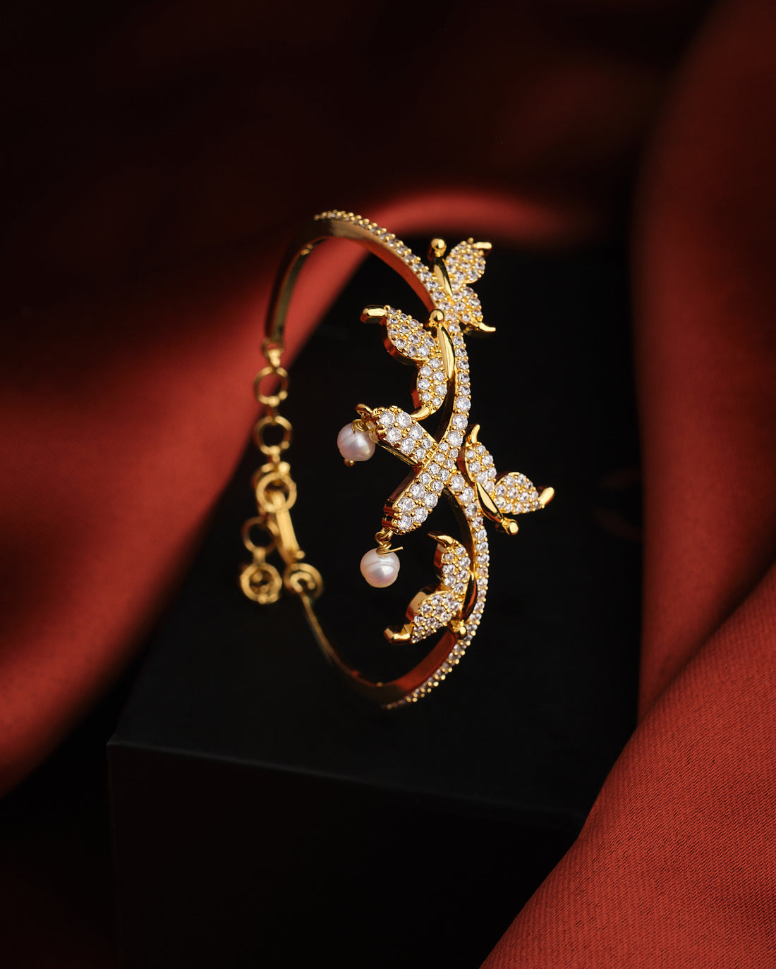 The Ensnaring Pearl Bangle by Chandrani Pearls is a vintage gold bracelet featuring leaf-shaped designs with small white stones and pearls, shown on a black surface with a rust-colored fabric backdrop.