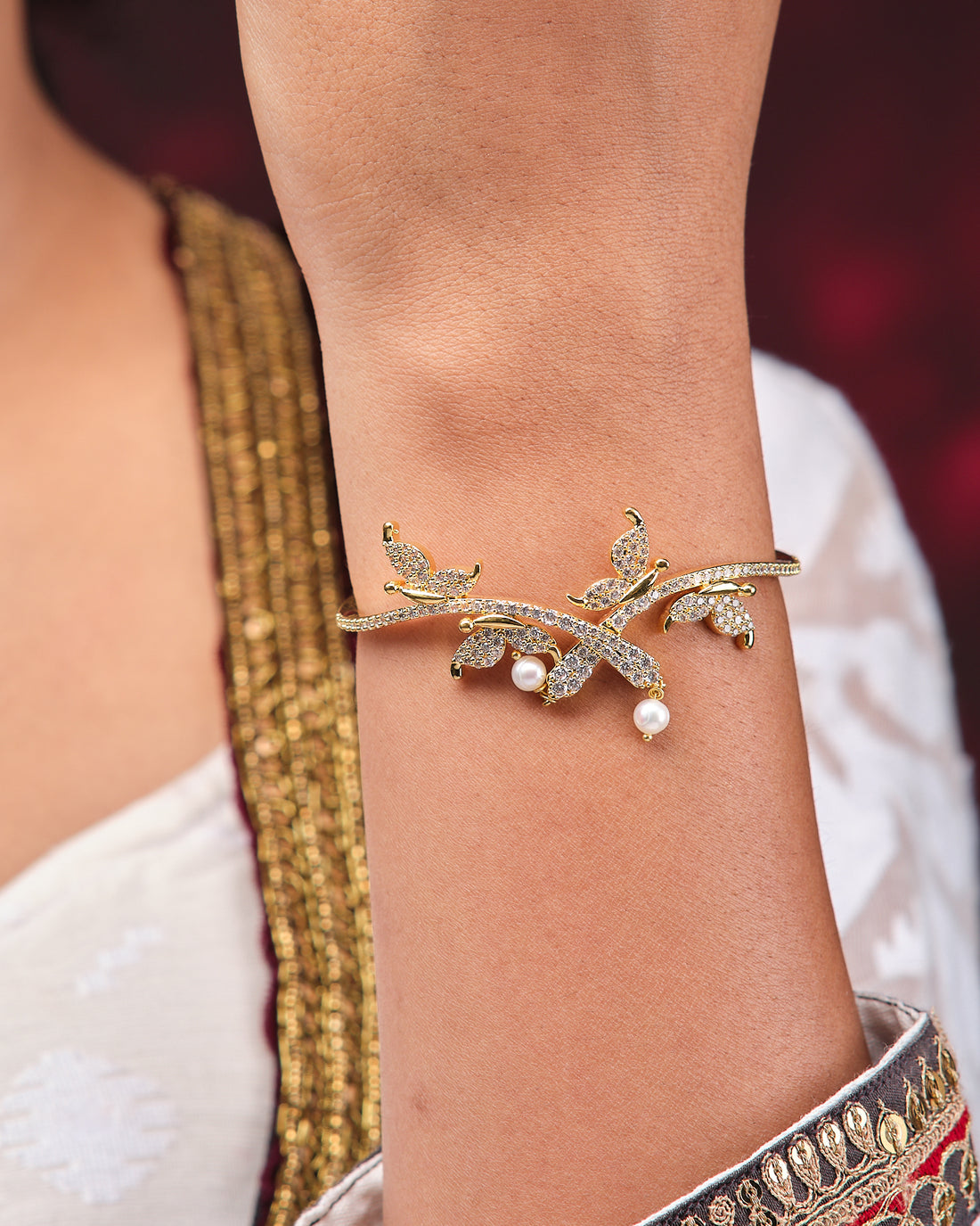 The Ensnaring Pearl Bangle by Chandrani Pearls is a vintage gold bracelet featuring leaf-shaped designs with small white stones and pearls, shown on a black surface with a rust-colored fabric backdrop.