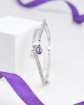 The Elegant Sterling Silver Bangle with Sparkling Cubic Zirconia by Chandrani Pearls features rows of shimmering stones, a teardrop purple gem, a pearl charm, and is displayed with a purple ribbon in the background.