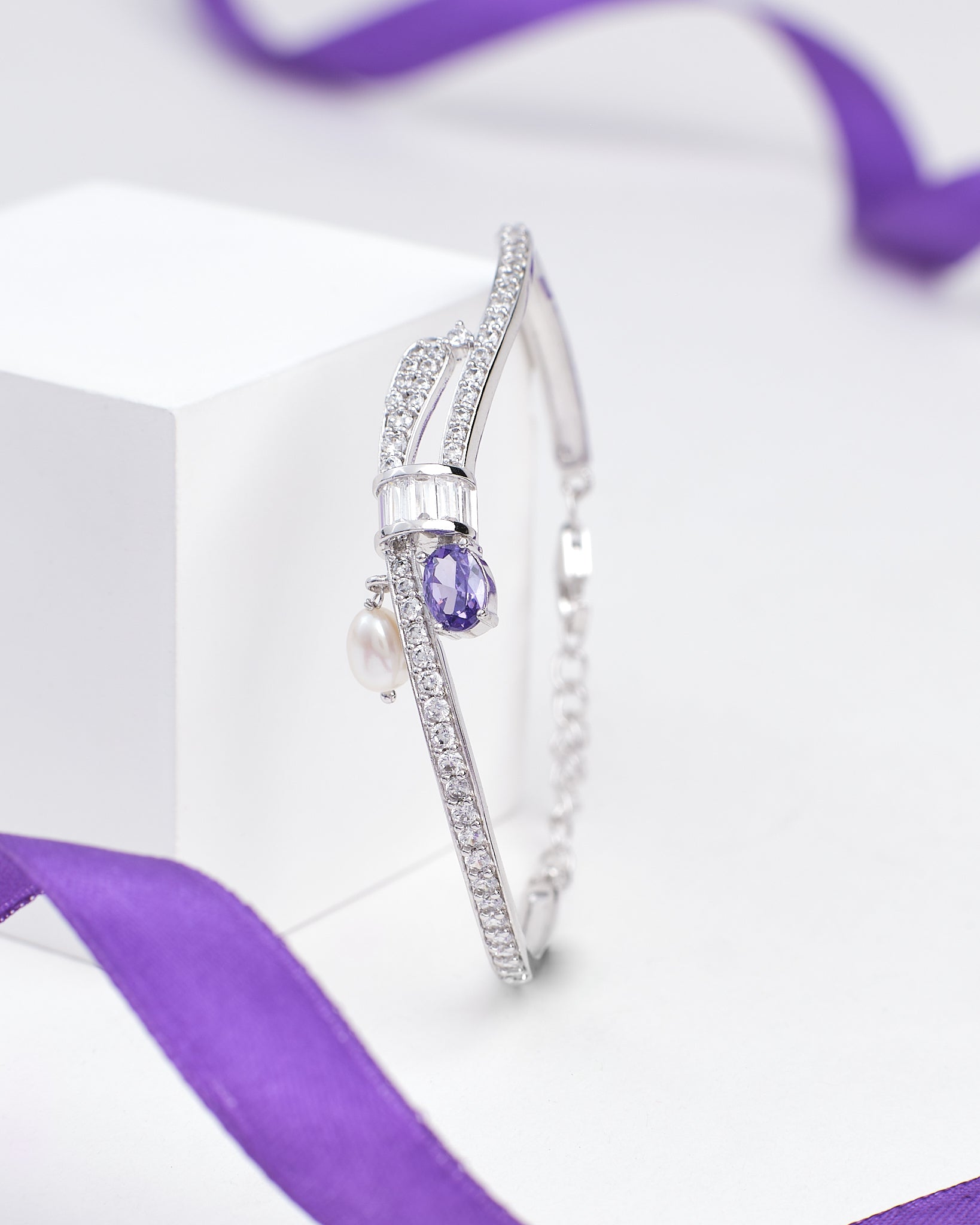 The Elegant Sterling Silver Bangle with Sparkling Cubic Zirconia by Chandrani Pearls features rows of shimmering stones, a teardrop purple gem, a pearl charm, and is displayed with a purple ribbon in the background.