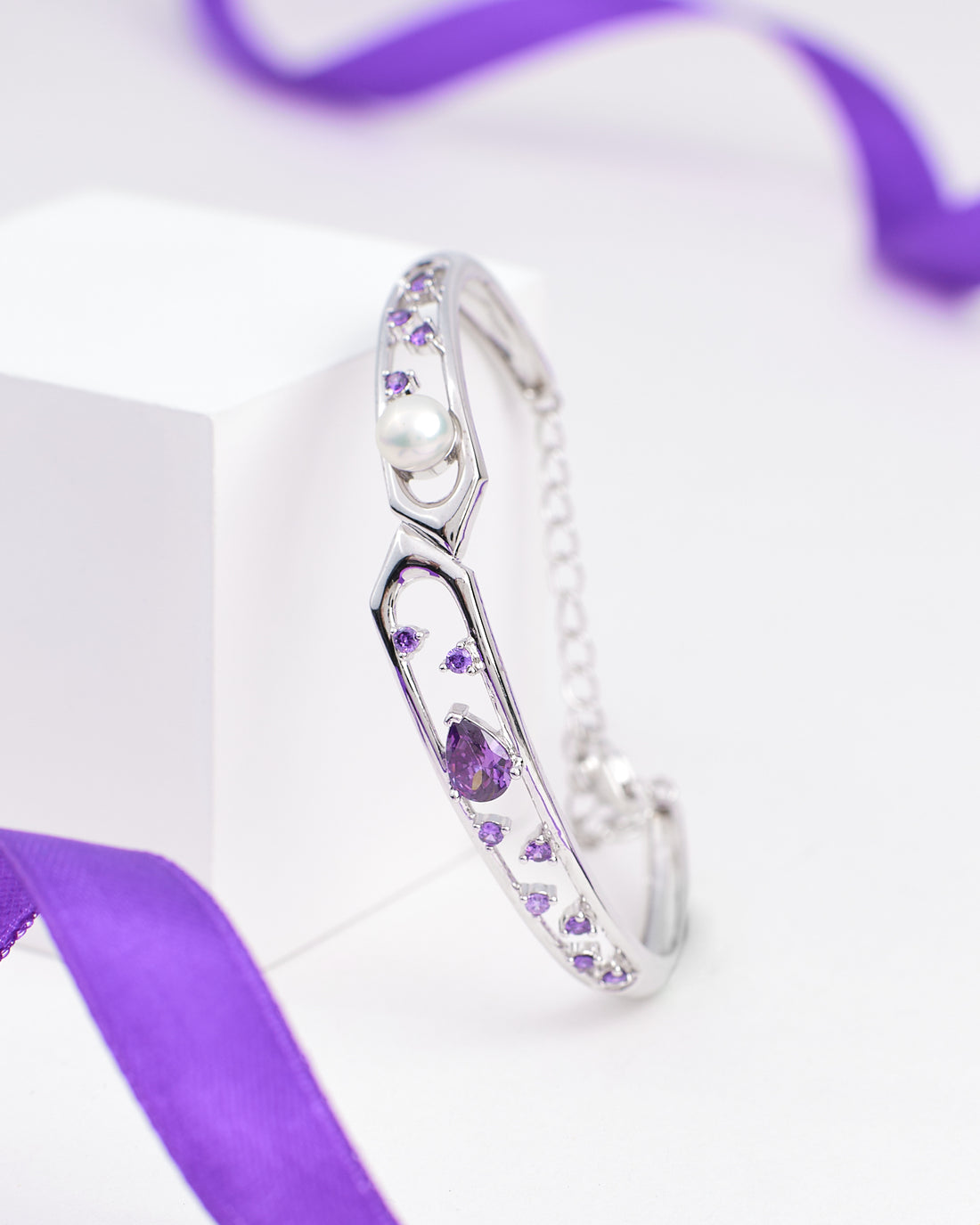 The Trendy Sterling Silver CZ Bangle by Chandrani Pearls, featuring purple gemstones, cubic zirconia, and a pearl, is displayed on a white block with a purple ribbon in the background.