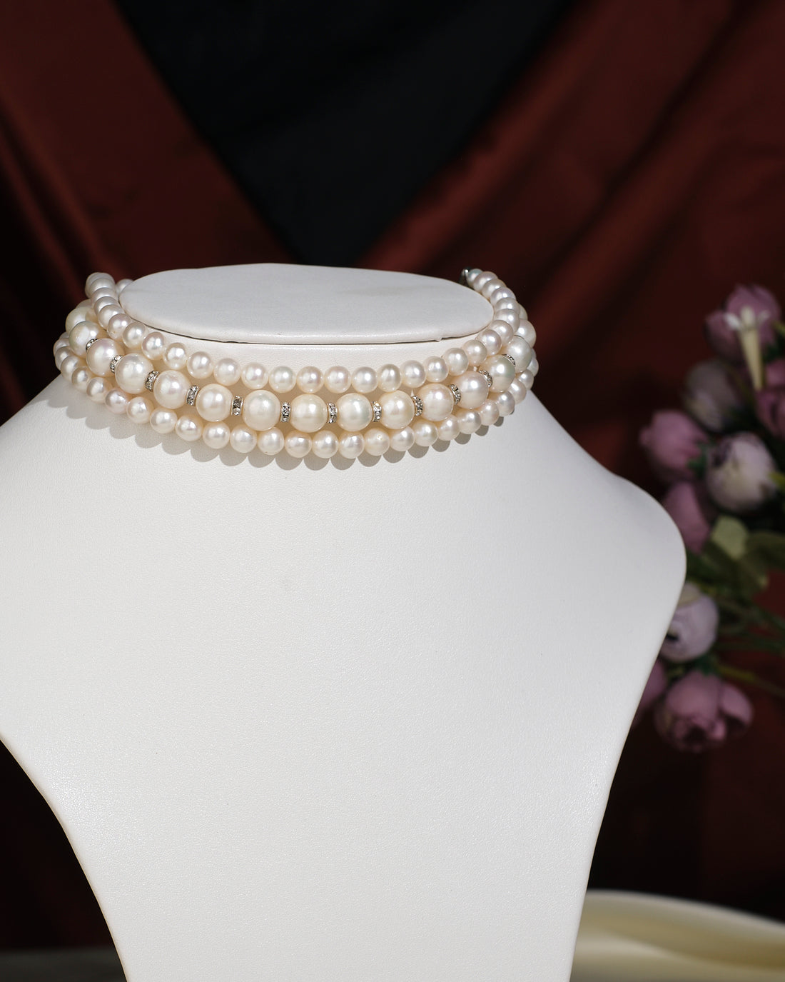 The Elegant Triple Line Choker by Chandrani Pearls India showcases refined glamour on a white jewelry stand, set against a softly blurred floral backdrop.