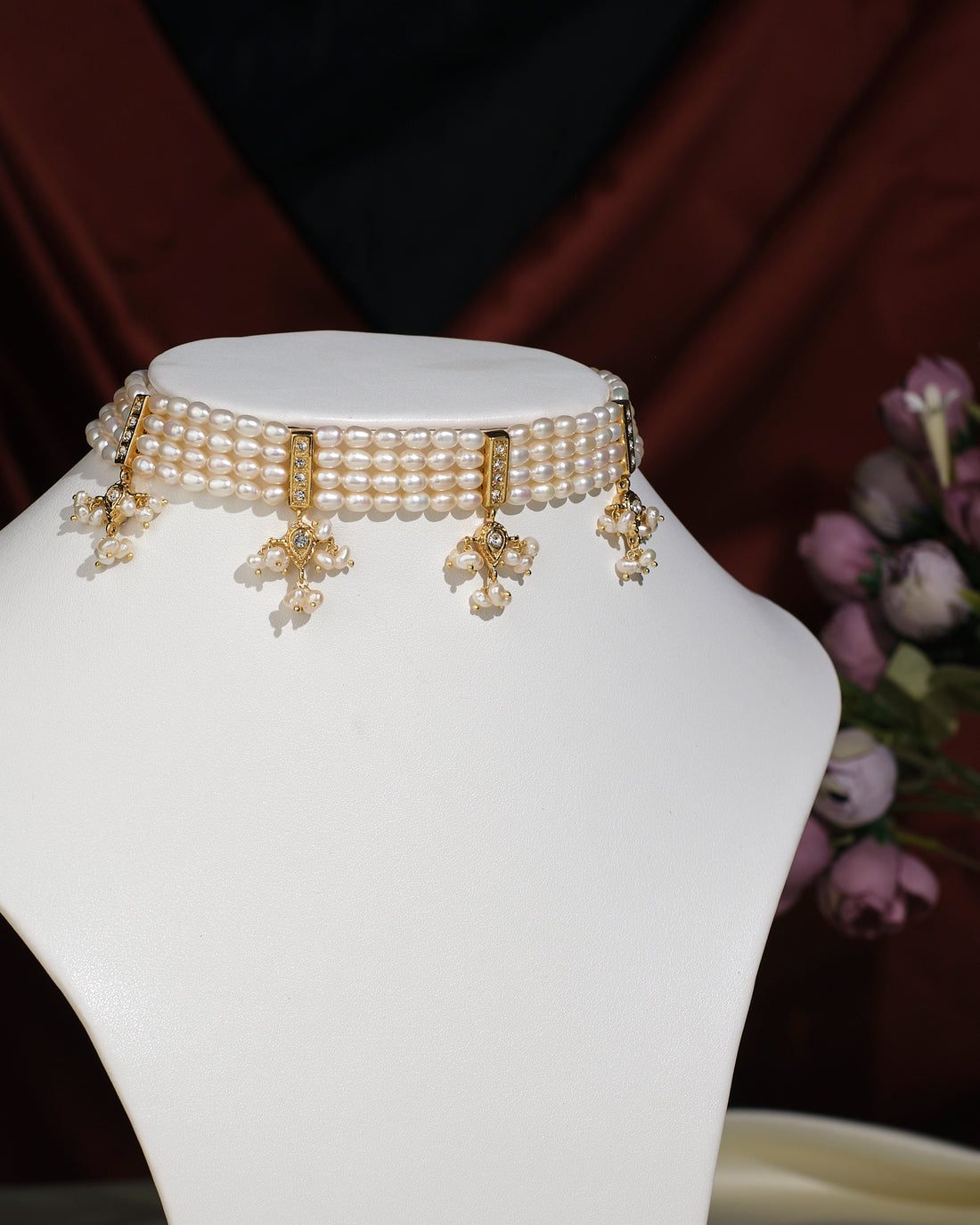 The Gorgeous 4 Line Pearl Choker by Chandrani Pearls India features gold and pearl floral pendants, exuding natural elegance on a white jewelry stand set against a dark background with purple flowers.