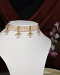 The Gorgeous 4 Line Pearl Choker by Chandrani Pearls India features gold and pearl floral pendants, exuding natural elegance on a white jewelry stand set against a dark background with purple flowers.