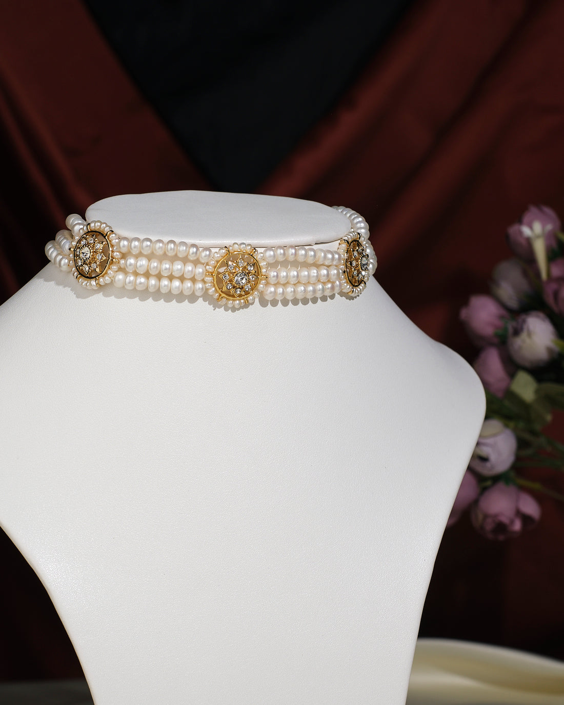 The Trendy 3 Line Pearl Choker by Chandrani Pearls India features gold and rhinestone floral accents, elegantly displayed on a white stand with a blurred flower arrangement in the background.