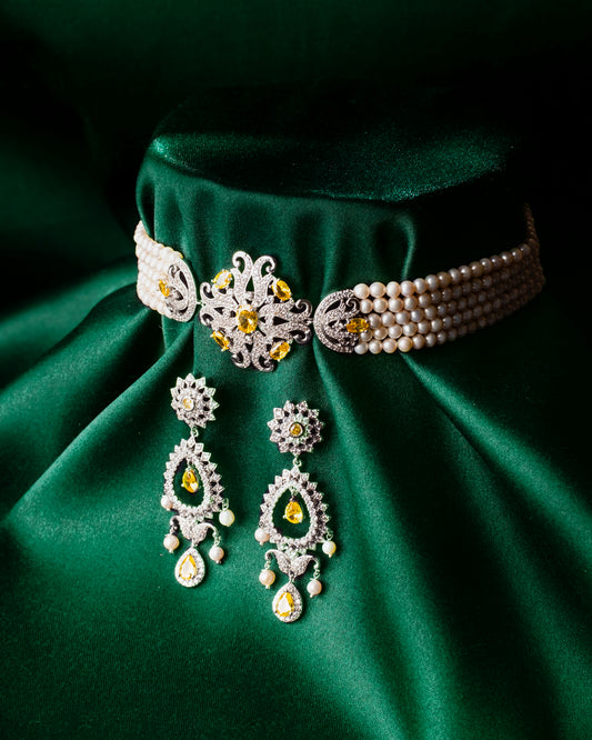 Displaying spectacularly on a green satin background, Chandrani Pearls' Deepa's Regal Flair Necklace Set includes a pearl choker with an ornate central piece featuring yellow gemstones. The set is beautifully complemented by matching drop earrings adorned with green, yellow, and white gems, exuding true regal flair.