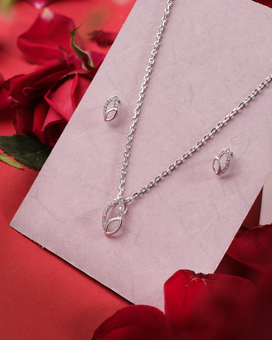 The V' Loop Leafy 925 Silver White Pendant Set by Chandrani Pearls India, with its elegant oval leaf design, is displayed on a pink card surrounded by red rose petals for a refined and sophisticated style.