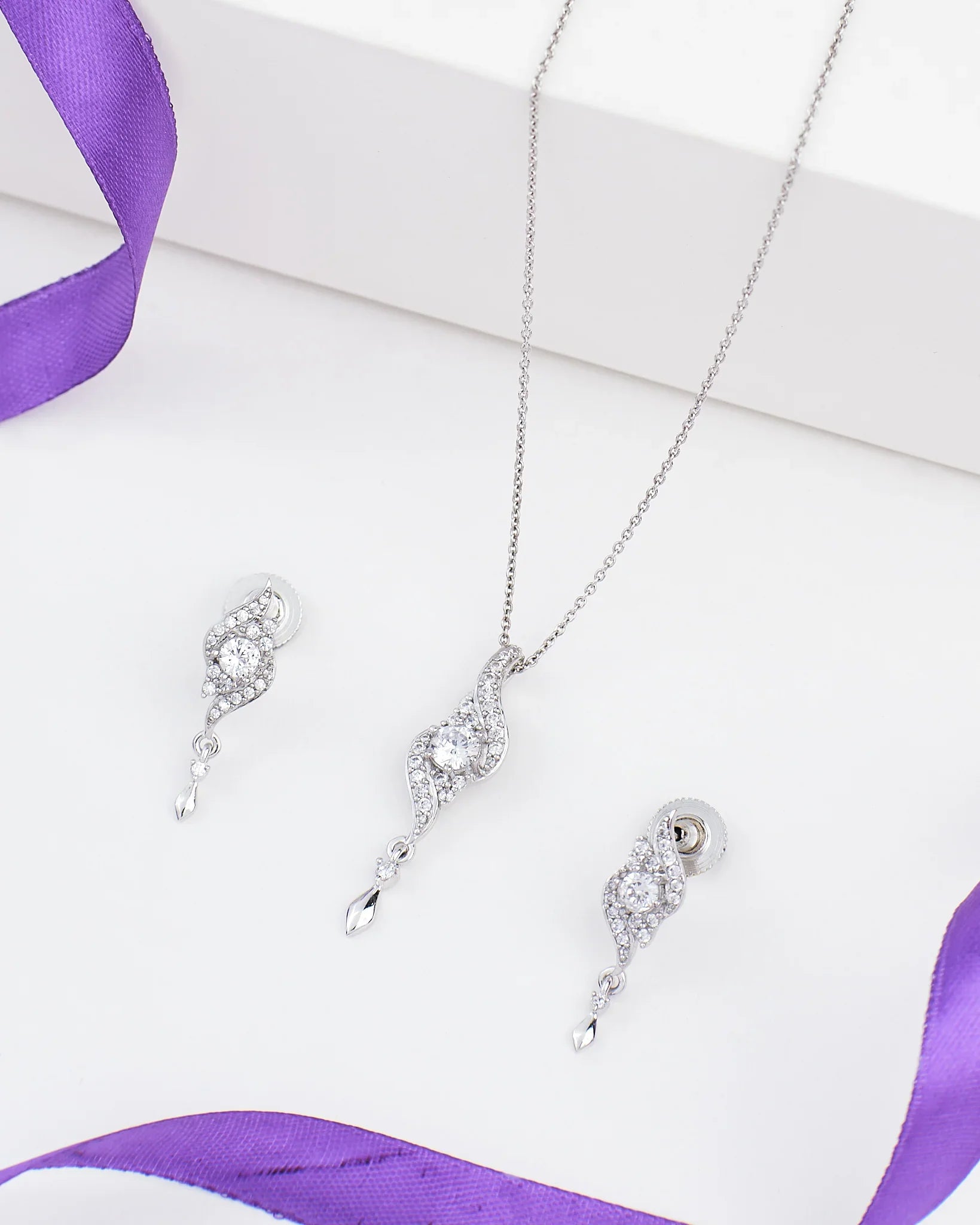 The Chandrani Pearls Floral Grace Pendant Set in sterling silver with CZ and pearl is displayed on a white surface next to a purple ribbon.