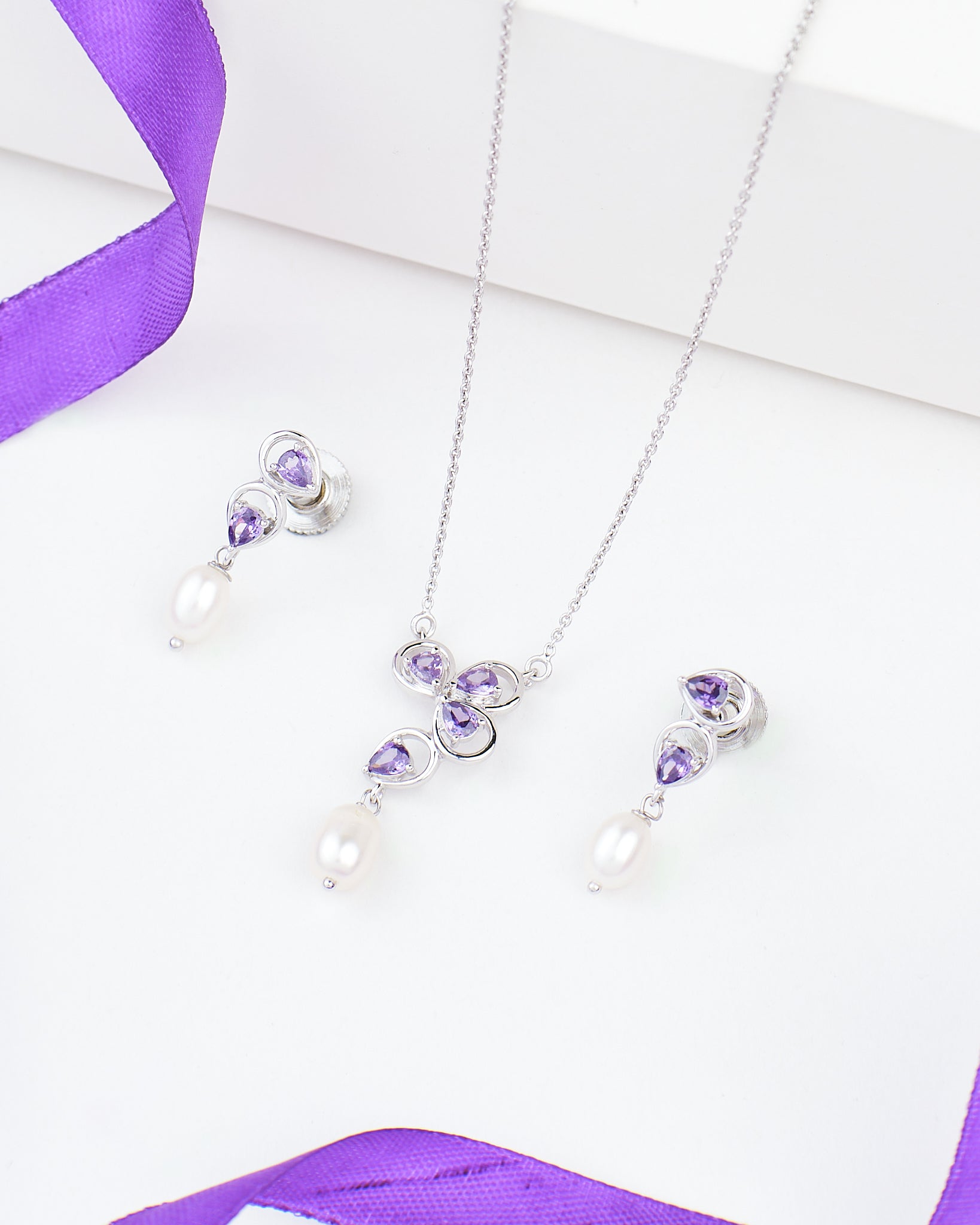 The Purple Celestial Spark Pendant Set by Chandrani Pearls features 925 silver with purple CZ, lustrous pearls, and comes with a matching necklace and earrings, displayed on a white background with purple ribbon accents.