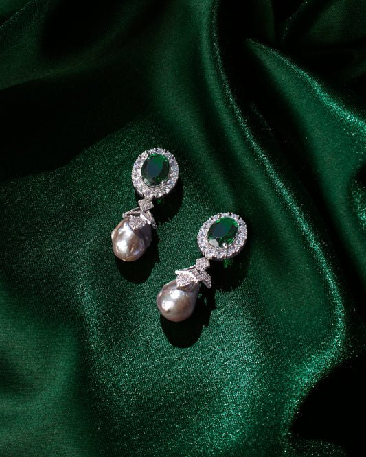 A pair of **Deepa's Emerald and Baroque Pearl Earrings** by **Chandrani Pearls**, displaying elegant green gemstones surrounded by smaller clear stones and featuring large pearls, set against a backdrop of green satin fabric.