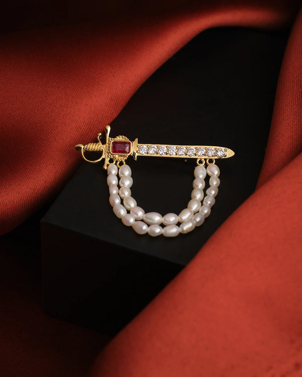 The Brawny Waves Pearl Brooch For Him by Chandrani Pearls features a gold sword shape, red gemstone, clear stones, and two strands of white pearls—an exquisite piece displayed on a dark surface with rust-colored fabric in the background.
