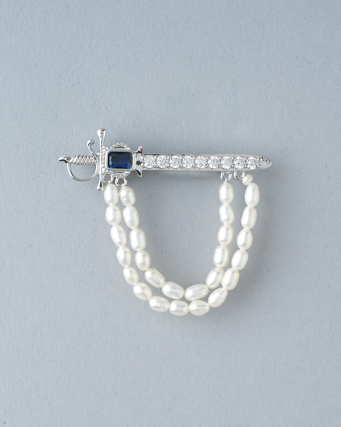 The Husky Gleam Pearl Brooch For Him by Chandrani Pearls, featuring a silver sword design with a black gemstone, rhinestones, and double pearl strands, exudes elegance atop a black surface with rust-colored fabric.