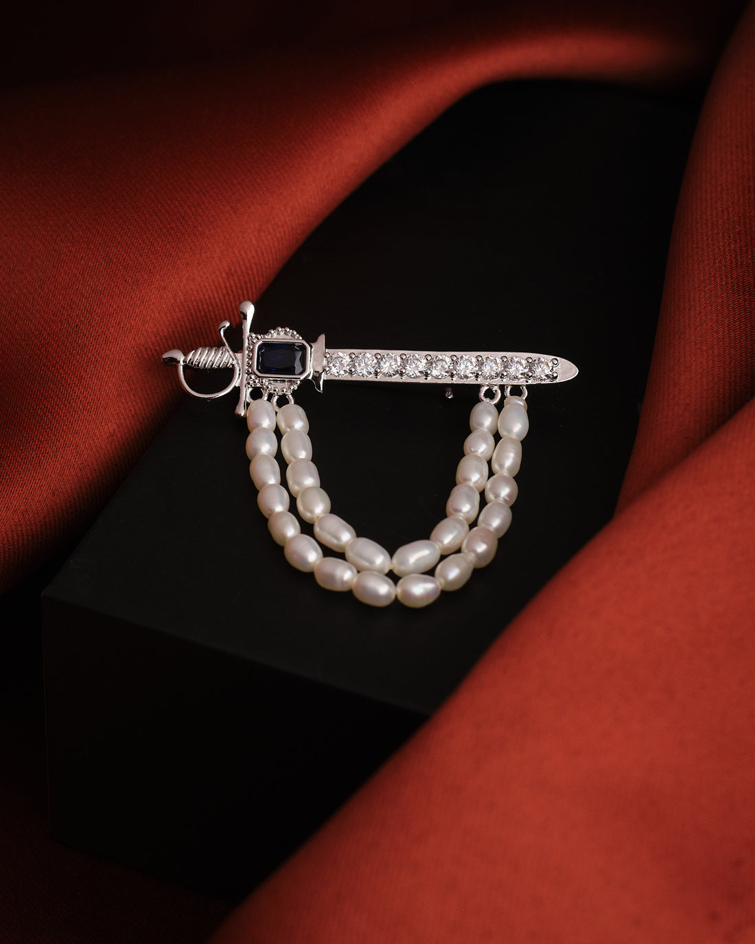 The Husky Gleam Pearl Brooch For Him by Chandrani Pearls, featuring a silver sword design with a black gemstone, rhinestones, and double pearl strands, exudes elegance atop a black surface with rust-colored fabric.