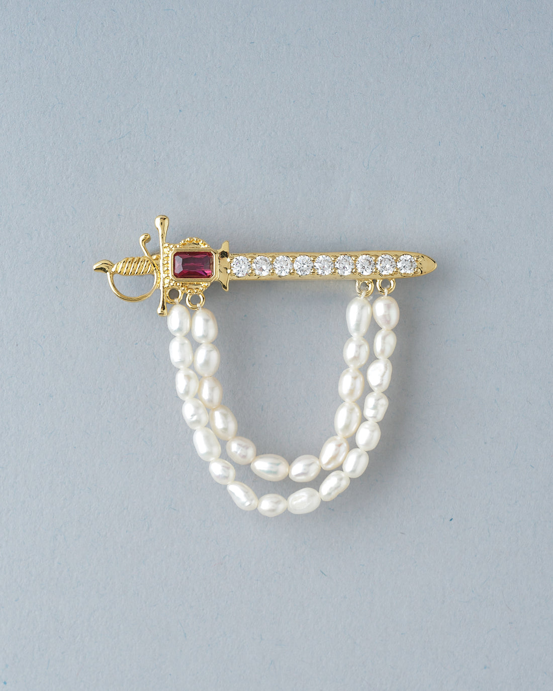 The Brawny Waves Pearl Brooch For Him by Chandrani Pearls features a gold sword shape, red gemstone, clear stones, and two strands of white pearls—an exquisite piece displayed on a dark surface with rust-colored fabric in the background.