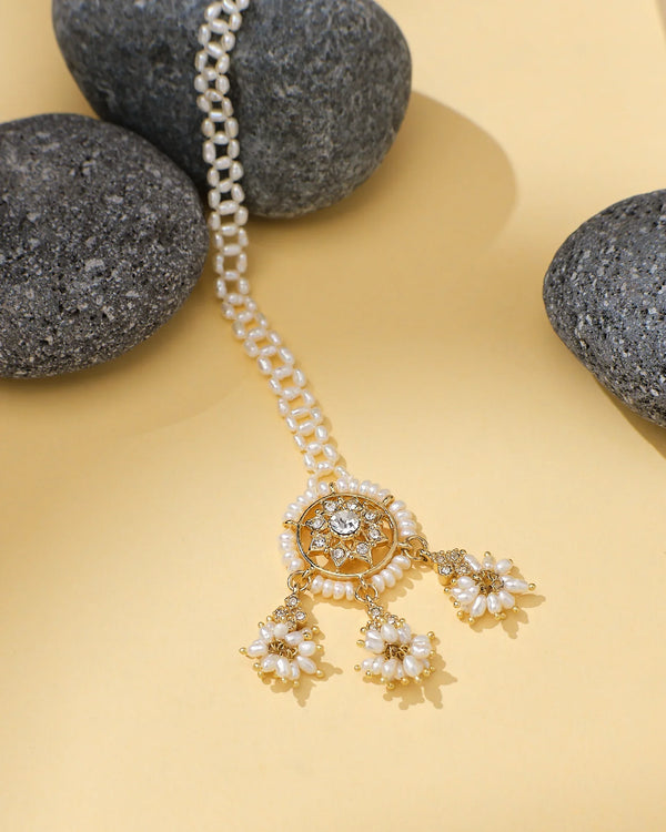 The Gorgeous Pearl CZ Tikli Timeless Bridal Grace by Chandrani Pearls, featuring a gold circular pendant with pearl clusters and a striking pearl centerpiece, is displayed on a yellow surface beside dark gray stones.