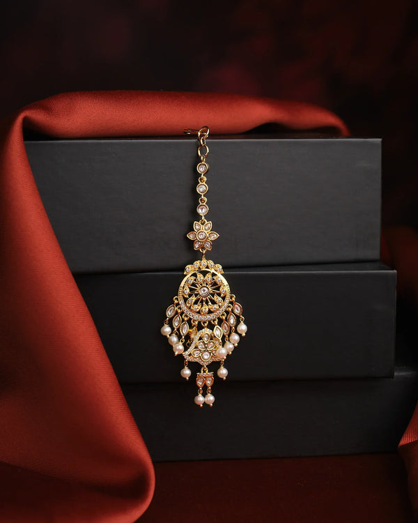 The Ethereal Grace Pearl Mang Tikka by Chandrani Pearls, adorned with pearls and kundan stones, is displayed on stacked black boxes and a red drape—an exquisite wedding accessory.