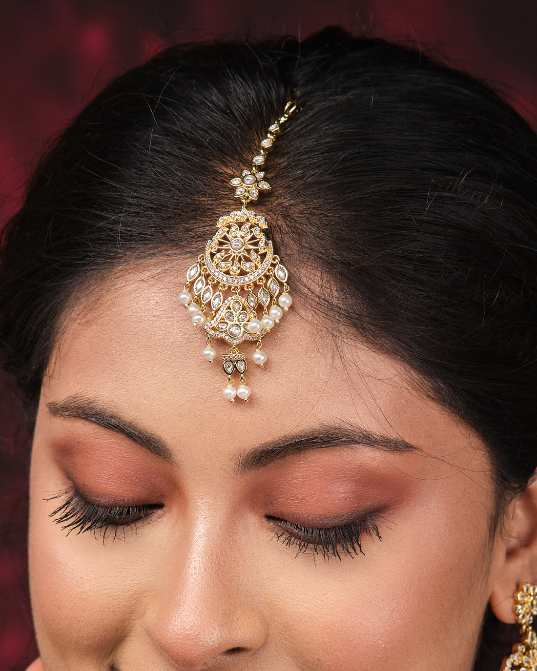 The Ethereal Grace Pearl Mang Tikka by Chandrani Pearls, adorned with pearls and kundan stones, is displayed on stacked black boxes and a red drape—an exquisite wedding accessory.