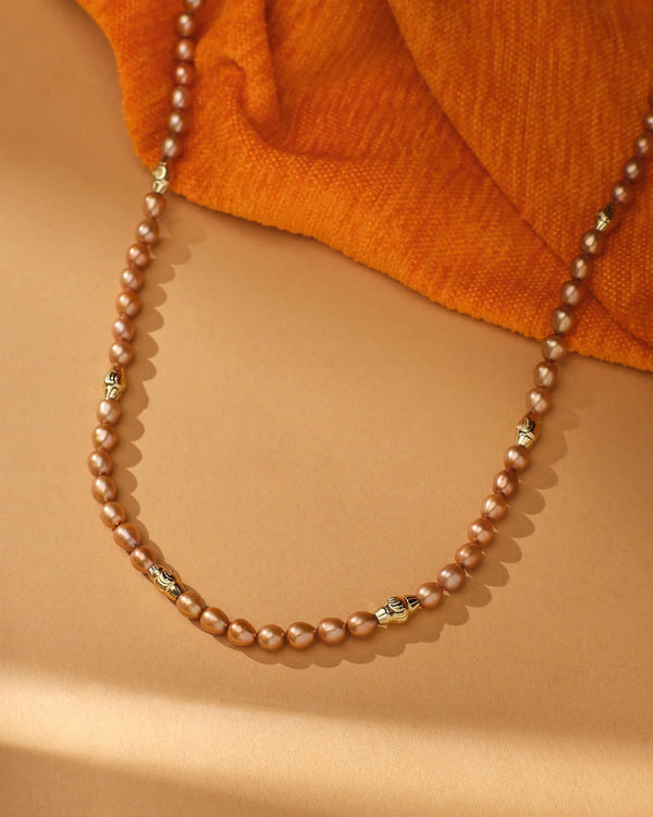 The Aurora Pearl Necklace by Chandrani Pearls, featuring brown pearls and gold accents, is showcased on a tan surface with orange textured fabric in the background.