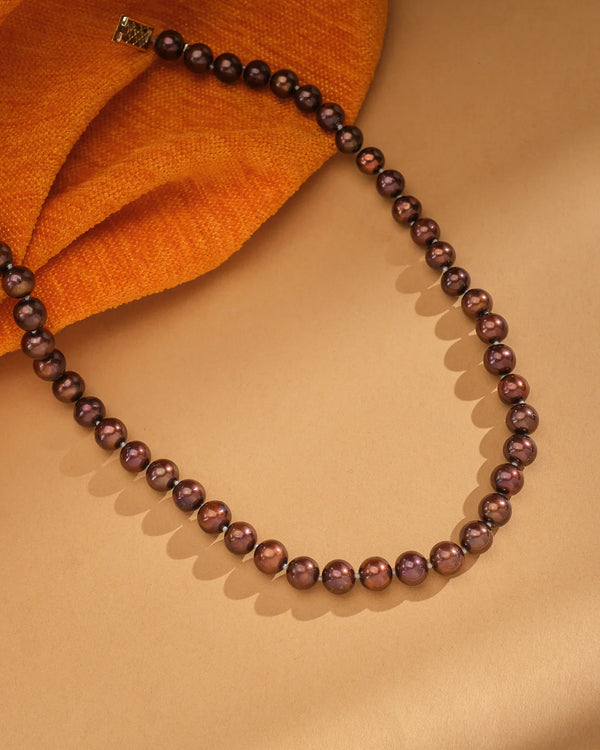 The Luna Pearl Round Necklace by Chandrani Pearls, featuring dark brown pearls and a gold clasp, rests on a tan surface beside orange textured fabric.