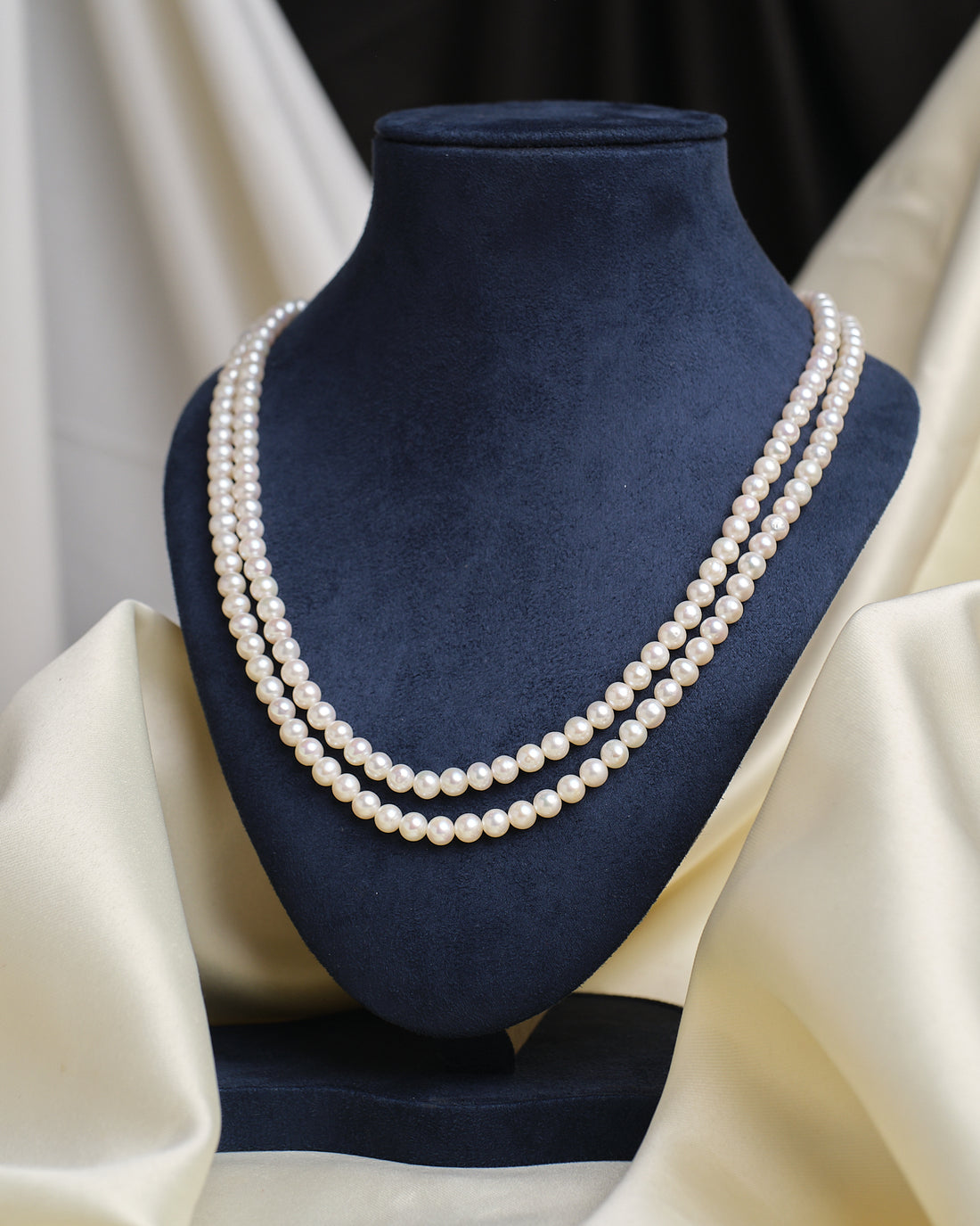 The Classic Single-Strand Pearl Necklace by Chandrani Pearls is displayed on a navy blue velvet bust, with cream-colored fabric draped in the background.