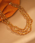 The Celeste Un-Shaped Gold Layered Necklace by Chandrani Pearls features three strands of irregular gold-colored beads with a clasp, displayed on a light brown surface beside orange textured fabric—a trendy choice for jewelry lovers.