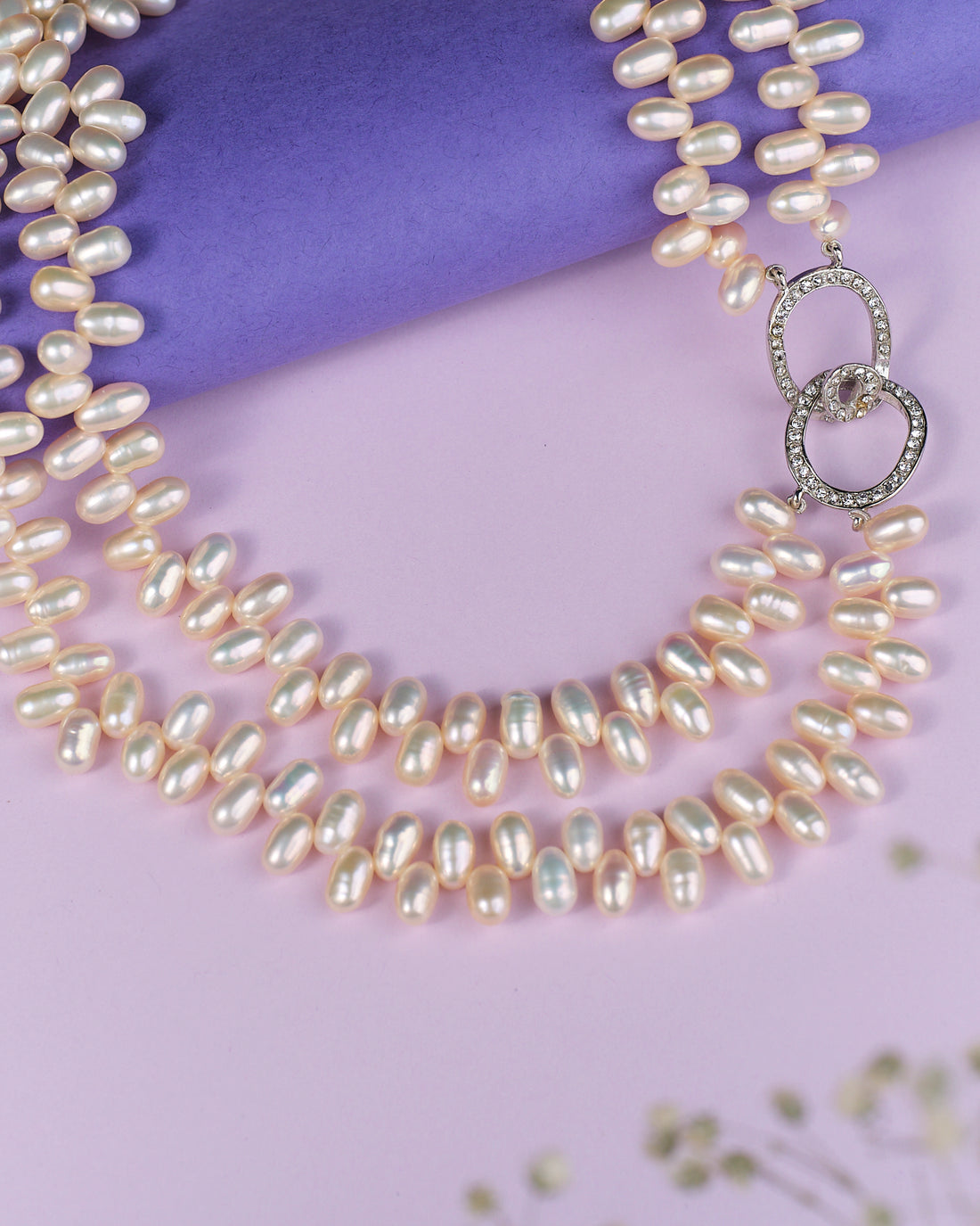 The Lavish Layers Of Elegance Pearl Necklace by Chandrani Pearls exudes sophistication as it rests gracefully on a light purple surface, with a deeper purple roll enhancing the background.