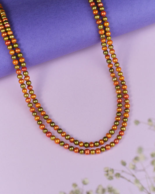 The Hiranya Multi Colour Pearl Necklace by Chandrani Pearls features two lustrous metallic bead strands in gold and copper hues, set against a purple and lavender backdrop with blurred floral details.