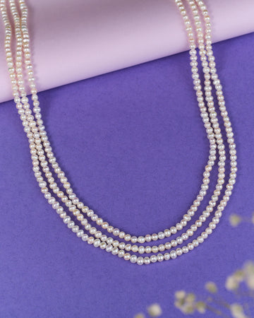 The Glistening White Peony Pearl Necklace by Chandrani Pearls exudes class and refinement, elegantly displayed on a purple surface with a soft pink roll in the background.