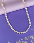 The Heavenly Pearl Necklace by Chandrani Pearls features elegant white pearls arranged in a circular form on a purple and pink background with delicate white flowers in the foreground.
