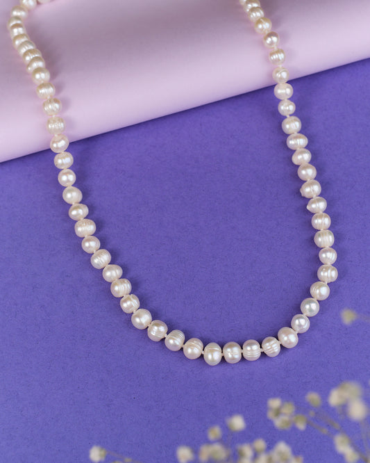 The Heavenly Pearl Necklace by Chandrani Pearls features elegant white pearls arranged in a circular form on a purple and pink background with delicate white flowers in the foreground.