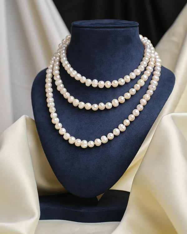 A navy blue velvet display stand showcases Chandrani Pearls' Colored Pearl Necklace, featuring three strands of white pearls, set against a cream and black fabric background.