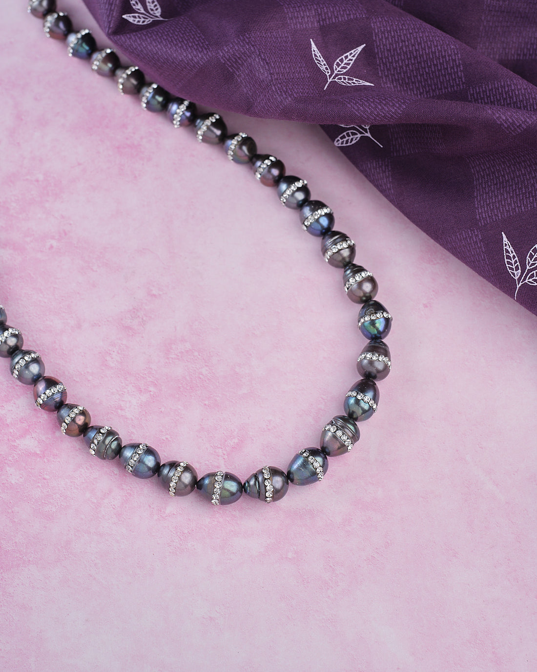 The Splashy Sleet Pearl Necklace by Chandrani Pearls, with patterned black and silver beads, is displayed on a pink textured surface next to a folded purple cloth adorned with white leaf designs.