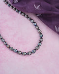 The Splashy Sleet Pearl Necklace by Chandrani Pearls, with patterned black and silver beads, is displayed on a pink textured surface next to a folded purple cloth adorned with white leaf designs.