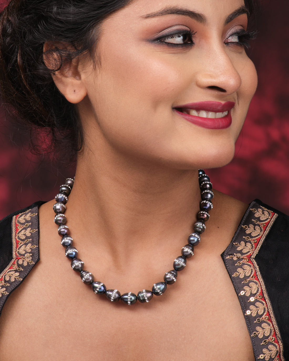 The Splashy Sleet Pearl Necklace by Chandrani Pearls, with patterned black and silver beads, is displayed on a pink textured surface next to a folded purple cloth adorned with white leaf designs.