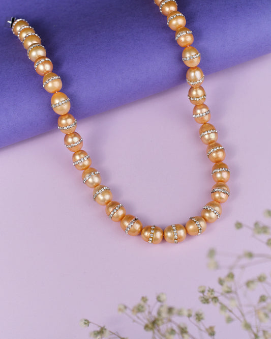 The Citizen Of The Globe Pearl Necklace by Chandrani Pearls features golden beads and rhinestone accents on a purple surface, captivating jewelry lovers. Delicately framed with small flowers, it's truly a stunning piece.