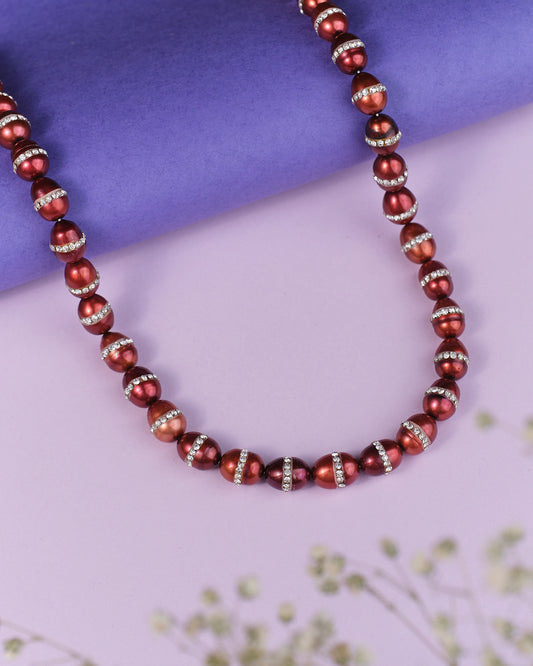 The Cherry On The Cake Pearl Necklace by Chandrani Pearls, featuring red pearls with silver spacers, elegantly rests on a blue and pink surface. Delicate white flowers enhance its allure, making it an ideal statement piece for any formal gown.