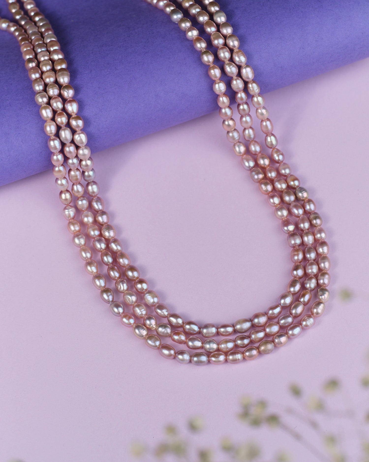 The Udagat - Blushing Rose Pearl Necklace by Chandrani Pearls is elegantly draped over a purple cylinder on a soft pink surface.