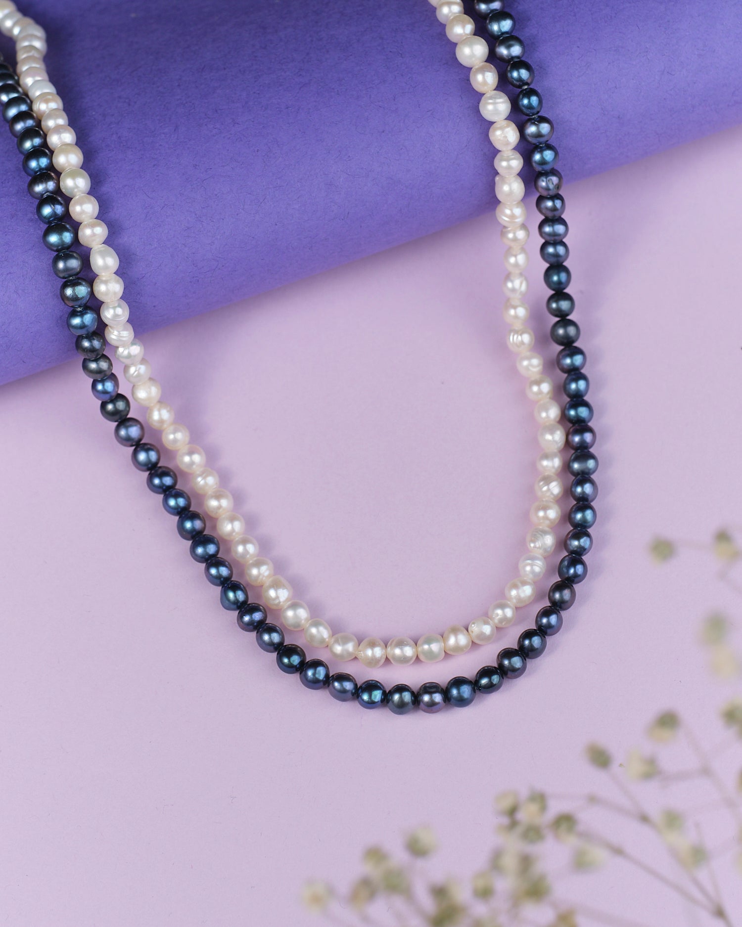 Two elegant Careen Axis Pearl Necklaces, one white and one black from Chandrani Pearls, rest on a purple and pink surface decorated with small white flowers. Perfect for elevating your wedding style.