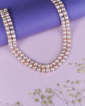 The Lavana-The Coast Memoirs Pearl Necklace by Chandrani Pearls, a luxurious double-stranded piece with pink and white pearls, gracefully drapes over a purple cylinder on a pink surface. Adorned with small white flowers, it is the perfect versatile accessory for any ensemble.